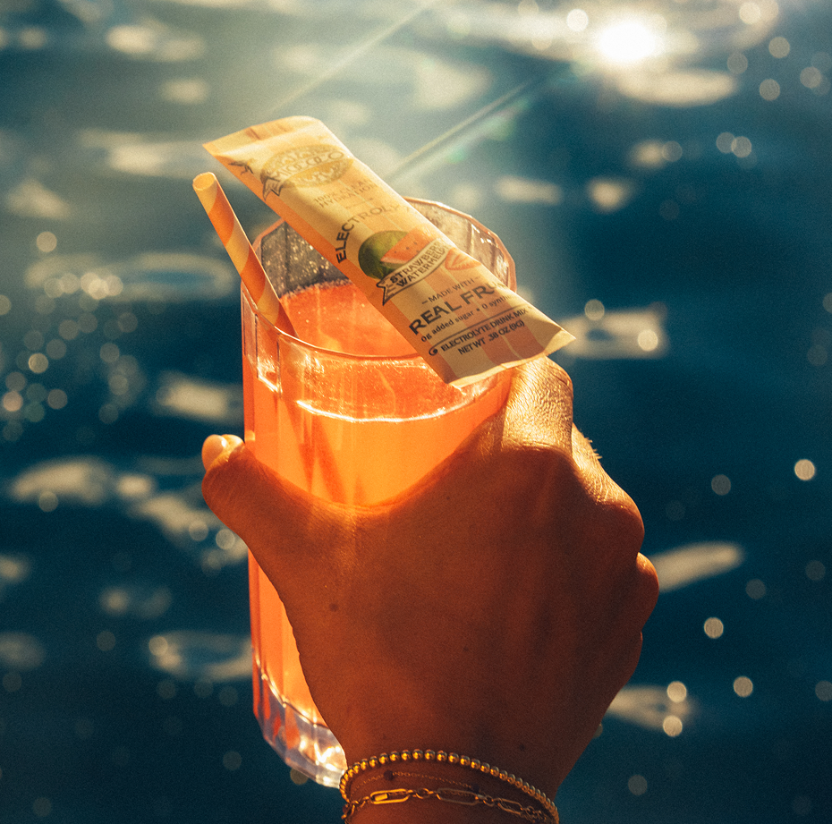 Hand holding an electrolyte beverage with a straw and a packet against a blue water background
