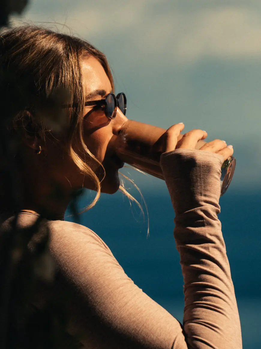 Woman wearing sunglasses with a blurred natural background drinking a chocolate protein shake.