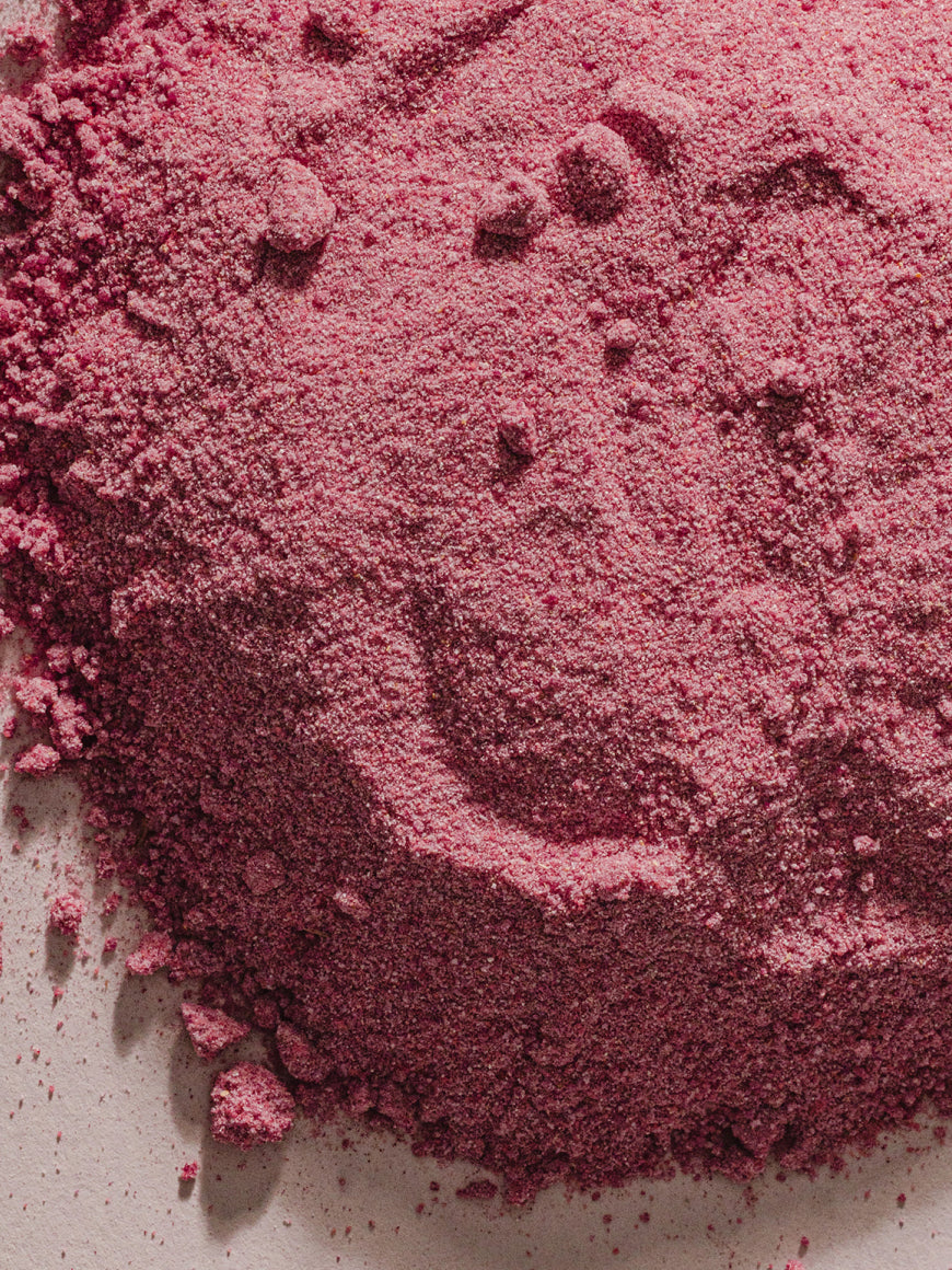 A close-up of loose, finely ground debloat powder in a dusty rose or mauve-pink shade, piled on a light neutral surface.