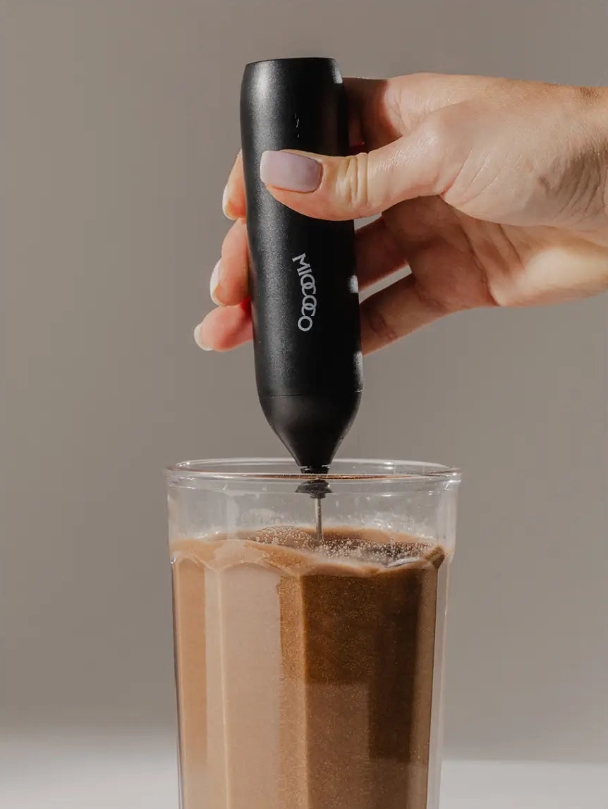A hand holding a black handheld frother branded 'MIOCOCO' above a glass of milk being frothed, against a plain taupe background.