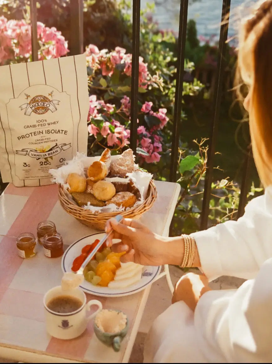 Person enjoying a breakfast scene mixing protein powder in their coffee with a basket of pastries, coffee, and fruit on a table outdoors.
