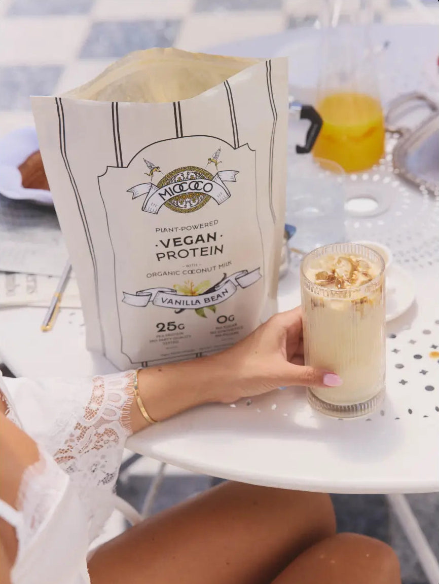 A person at a white table holds a glass of vanilla bean iced coffee next to a large bag of Micococo Vegan Protein powder.